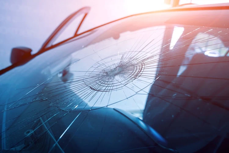 A car with a cracked windshield, showing a circular impact point radiating cracks outward. The car door is open, and sunlight filters through, illuminating the damage. The background is blurred, focusing on the windshield.