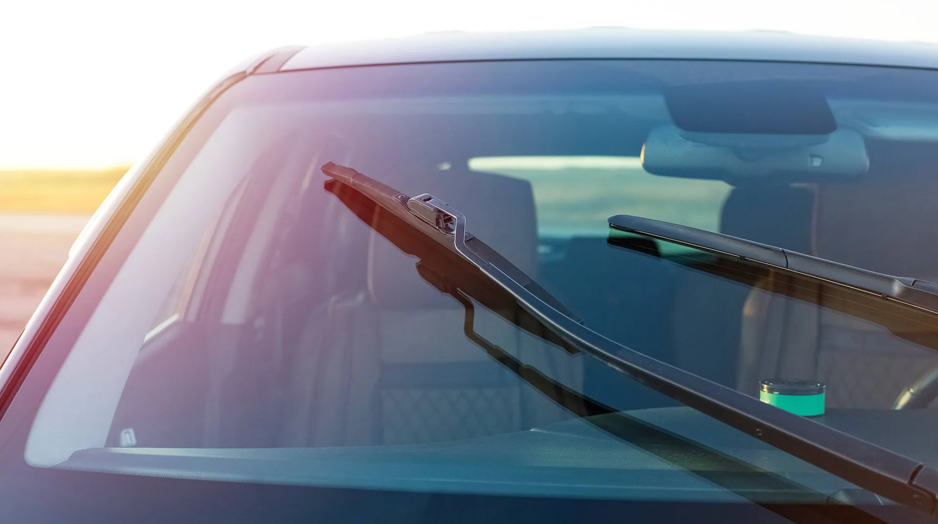 Close-up of a car windshield with two raised wipers, sunlight reflecting on the glass, part of the interior visible, and a bright, blurred outdoor background.