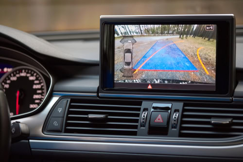 Dashboard view of a car’s rearview camera display showing parking guidelines with yellow and blue marked areas, with controls and part of the speedometer visible in the foreground.