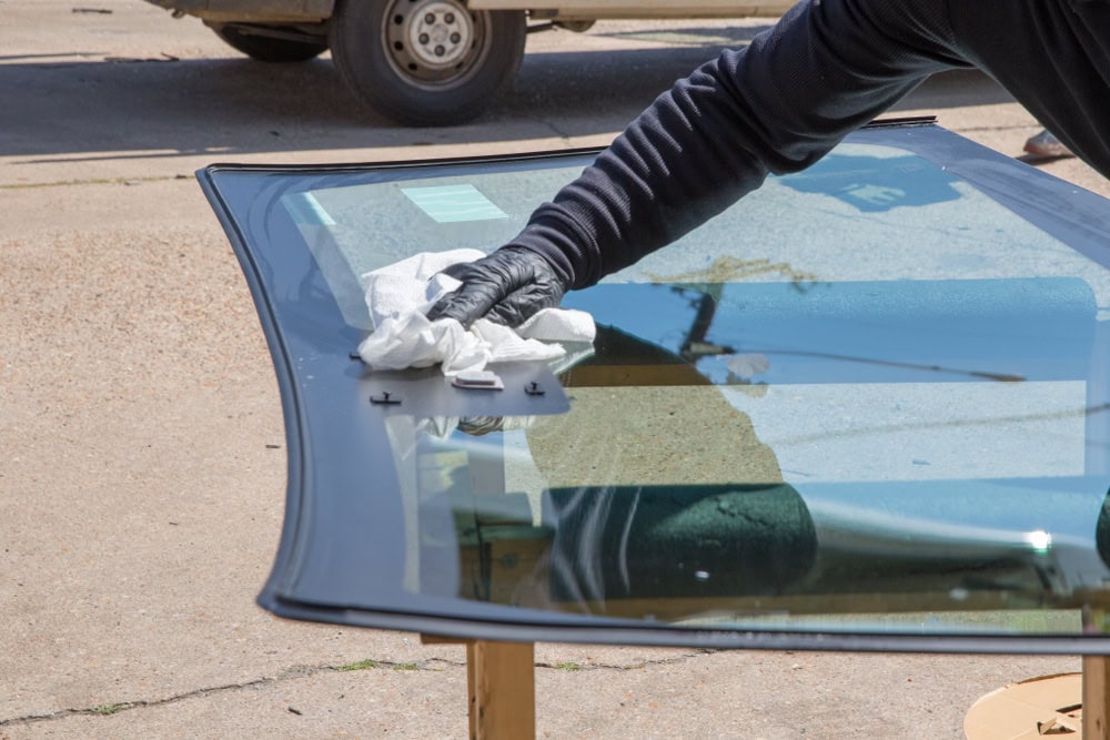 A person wearing a black glove wipes a large piece of curved glass, possibly an automobile windshield, with a white cloth outdoors on a sunny day.