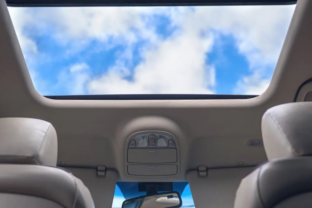View from inside a car looking up through an open sunroof, showing clear blue sky with scattered clouds. The car’s interior ceiling and headrests are also visible.