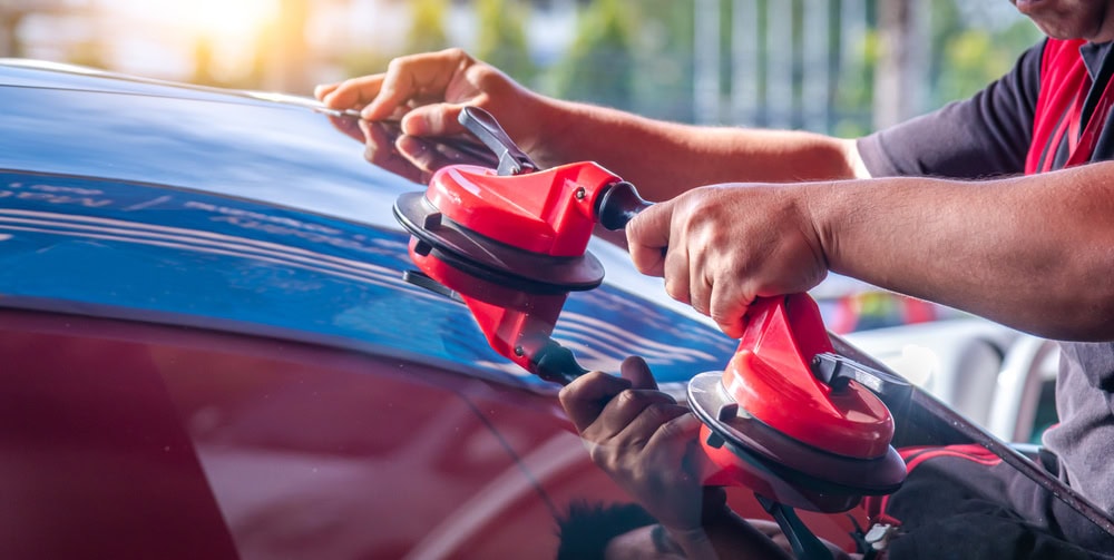 A person uses red suction tools to install or remove a car windshield, with sunlight reflecting off the glass surface.