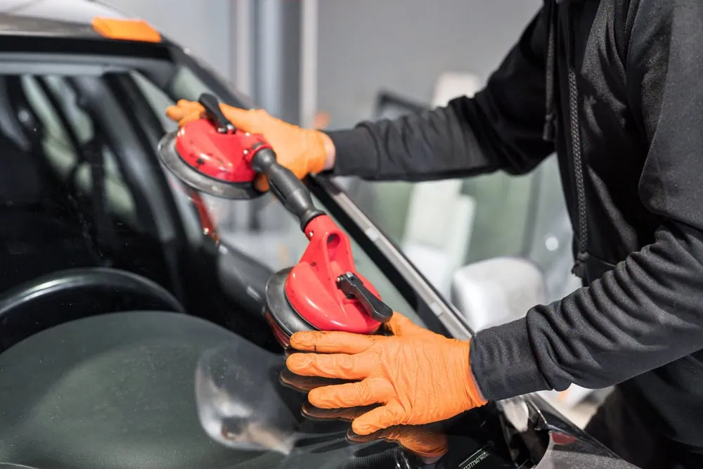 A person wearing orange gloves uses a red suction tool to carefully install or remove a windshield from a car.