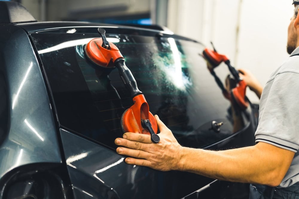 A person installs a rear windshield on a car using large red suction cups to carefully position the glass. The scene takes place in a workshop setting.