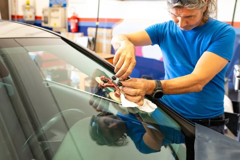A person wearing a blue shirt repairs a small crack or chip on a car windshield using a specialized tool inside an auto workshop.
