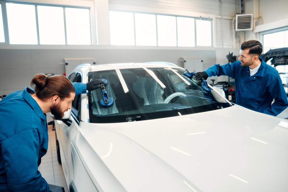 Two auto mechanics in blue uniforms work together inside a brightly lit garage to install or adjust the windshield of a white car, using large suction cup tools to lift and position the glass.