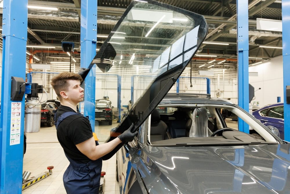 A mechanic wearing gloves and overalls installs or removes a windshield from a gray car inside a brightly lit auto repair shop.
