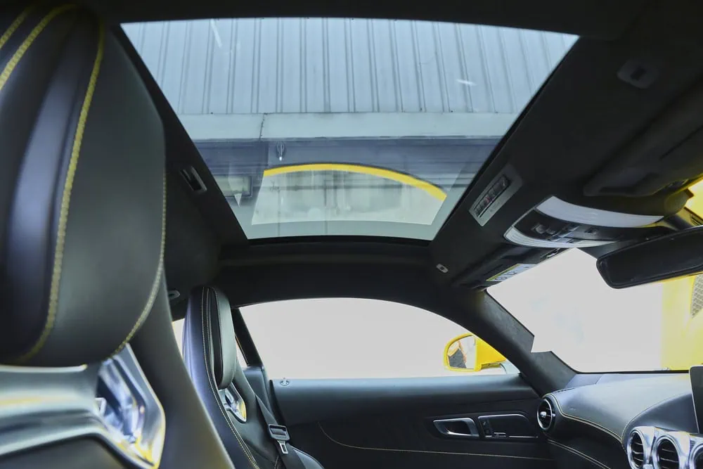 View from inside a luxury car with black leather and yellow stitching, showing a large sunroof, part of the dashboard, and a bright yellow exterior seen through the window.