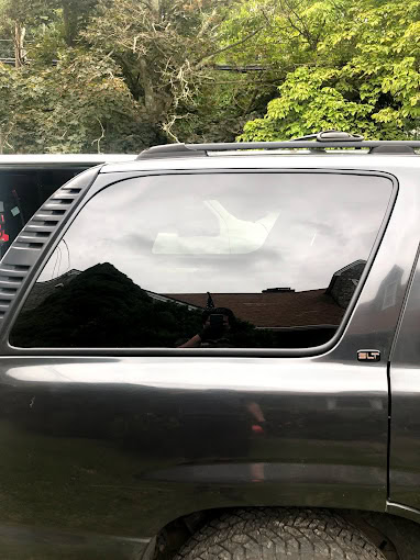 A shiny black SUV with tinted windows reflects nearby trees and a house. The vehicle is parked closely to another dark-colored car. Green foliage is visible in the background.