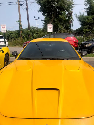 A bright yellow sports car is parked in a lot. The car's front and hood are visible. In the background, there are other cars, trees, and signs that read "Reserved Parking." The sky is clear, indicating a sunny day.