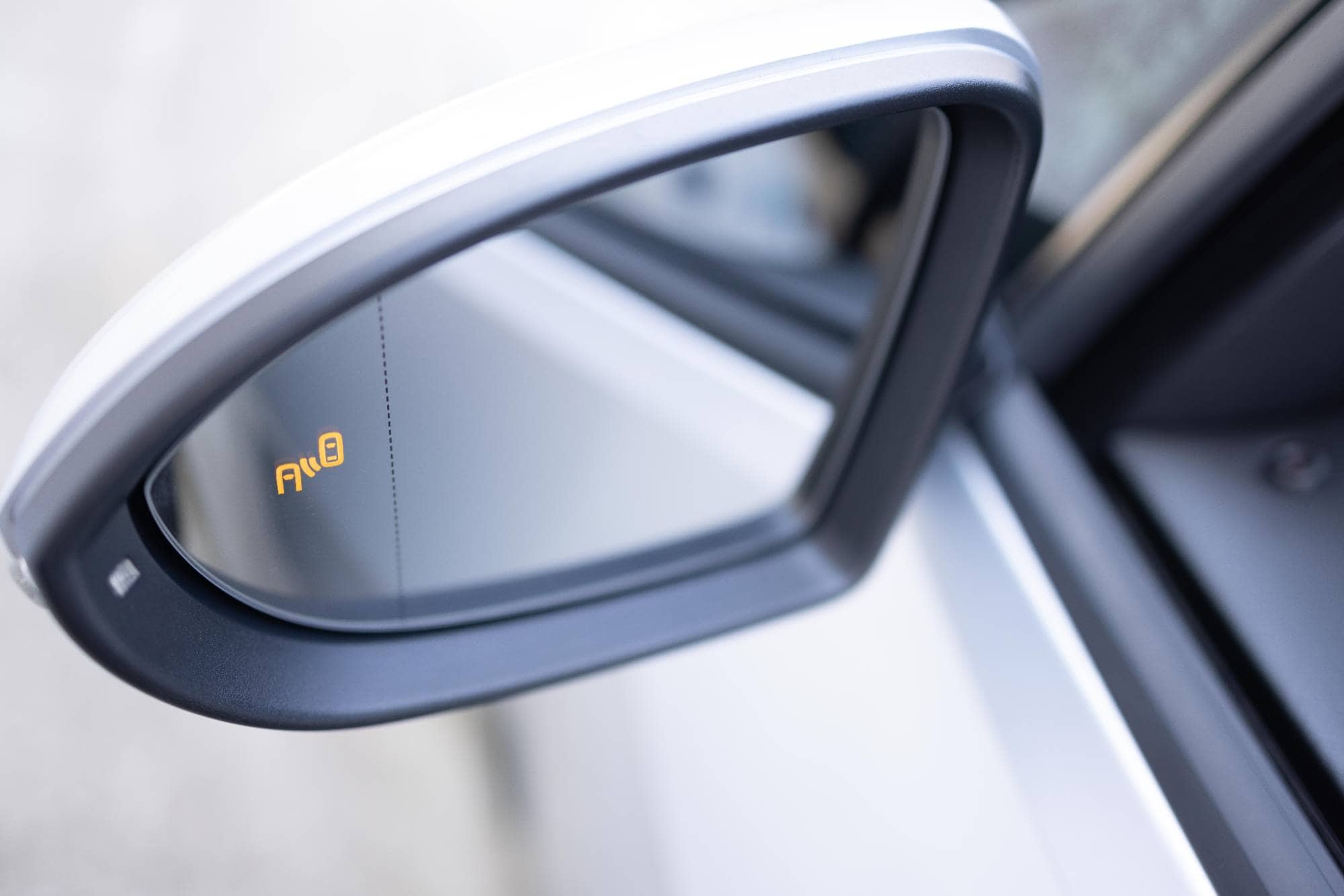 Close-up of a car’s side mirror with a glowing yellow blind spot warning icon, indicating a nearby vehicle in the driver’s blind spot, with part of the car door and exterior body visible.