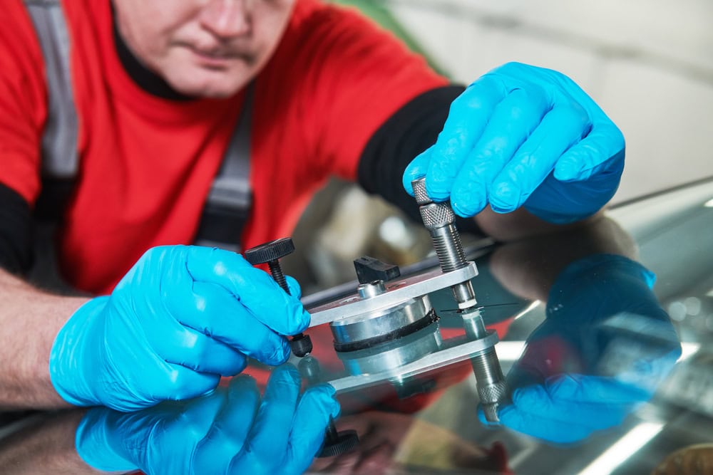 A person wearing blue protective gloves and a red shirt uses a specialized windshield repair tool to fix a crack or chip in a car’s glass, with the focus on their hands and the device pressed against the windshield surface.