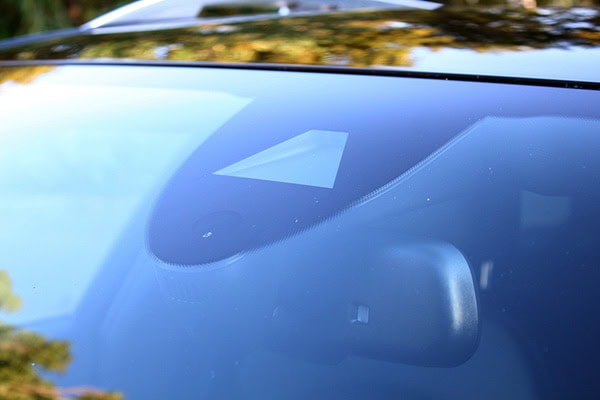 Close-up of a car windshield showing a small, triangular camera or sensor mounted near the top center, commonly used for driver assistance or safety features. Reflections of trees can be seen on the glass.