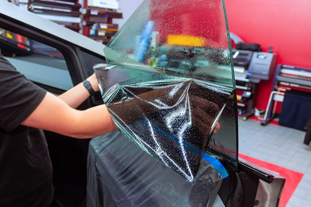 A person applies tinted film to a car window in a workshop, carefully smoothing the material over the glass to prevent bubbles. Various tools and equipment are visible in the background.