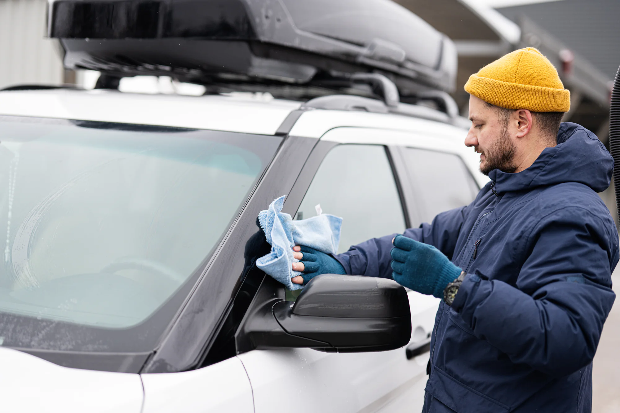 A man in a blue jacket, yellow beanie, and gloves cleans the side mirror of a white SUV with a blue cloth and spray bottle. The car, featuring a roof cargo box, is parked outdoors—Mobile Repair & Replacement Suffolk County, NY.