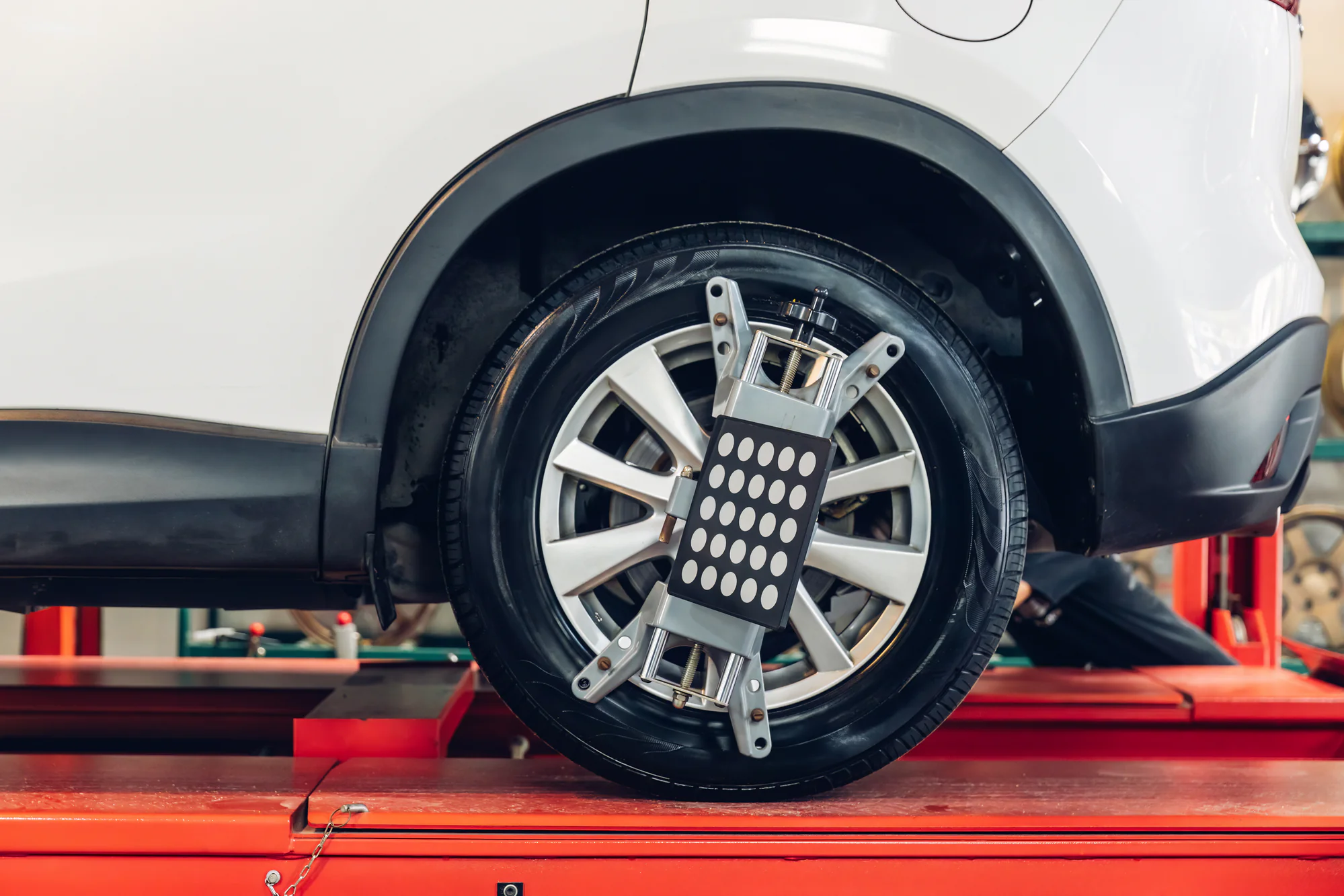 A close-up of a car tire with a wheel alignment tool attached, on a red platform in an auto repair shop. The vehicle appears to be undergoing maintenance or alignment service.