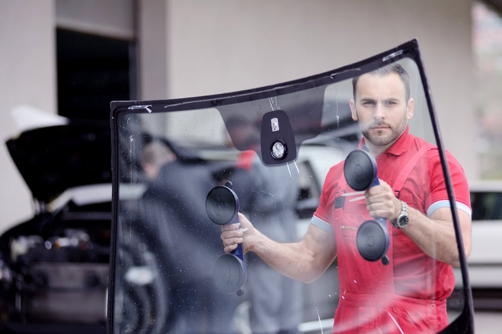 A man in a red uniform uses suction cup tools to hold a large car windshield—Mobile Repair & Replacement Suffolk County, NY. Behind him, a car with an open hood and another person blurred behind the glass are visible.